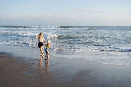 Foto de Madre con hija se divierten en la playa con arena negra en Bali, corriendo en las olas, disfrutar de la vida y viajar - Imagen libre de derechos