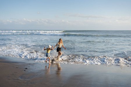 Téléchargez les photos : Mère avec sa fille s'amuser à la plage avec du sable noir à Bali, courir sur les vagues, profiter de la vie et voyager - en image libre de droit