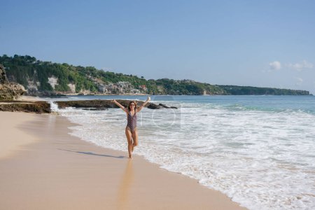 Téléchargez les photos : Femme gaie en maillot de bain en cours d'exécution dans l'océan mousseux atr plage de sable dans la journée ensoleillée. - en image libre de droit