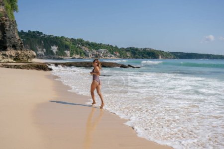 Foto de Alegre hembra en traje de baño corriendo en el océano espumoso atr playa de arena en el día soleado. - Imagen libre de derechos