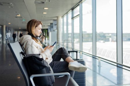 Foto de La mujer usa teléfonos inteligentes y auriculares mientras espera su vuelo, estudiante escuchando música o podcast en el aeropuerto mientras espera un vuelo. - Imagen libre de derechos