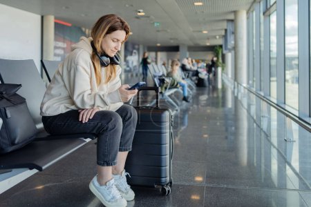 Foto de La mujer usa teléfonos inteligentes y auriculares mientras espera su vuelo, estudiante escuchando música o podcast en el aeropuerto mientras espera un vuelo. - Imagen libre de derechos