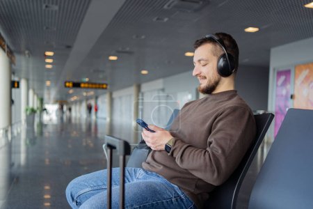 Foto de El joven está esperando un vuelo en el aeropuerto. Guy está escuchando música desde auriculares y usando un teléfono inteligente. - Imagen libre de derechos