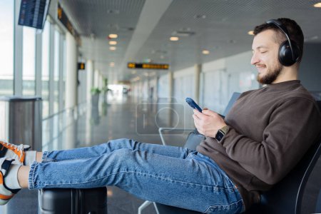 Téléchargez les photos : Le jeune homme attend le vol à l'aéroport. Guy écoute la musique des écouteurs et utilise un smartphone. - en image libre de droit