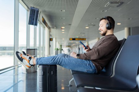 Téléchargez les photos : Le jeune homme attend le vol à l'aéroport. Guy écoute la musique des écouteurs et utilise un smartphone. - en image libre de droit