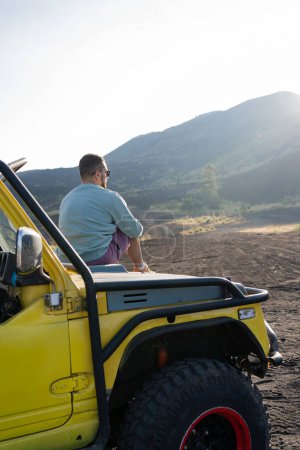 Foto de Hombre disfrutando de hermosos paisajes rocosos, sentado en el capó del coche en el borde de la carretera. Viajar por el valle volcánico de Bali. - Imagen libre de derechos