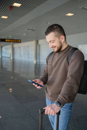 Téléchargez les photos : Portrait de l'homme joyeux se détendre à l'aéroport avec sac et téléphone mobile, gars en attente de vol en utilisant le téléphone mobile communiquer dans les médias sociaux, la recherche d'informations en ligne. - en image libre de droit