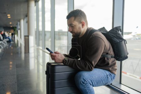 Photo for Portrait of cheerful man relaxing at airport with bag and mobile phone, guy waiting for flight using mobile phone communicating in social media, searching information online. - Royalty Free Image