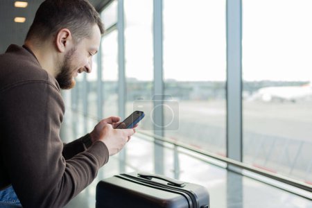 Photo for Portrait of cheerful man relaxing at airport with bag and mobile phone, guy waiting for flight using mobile phone communicating in social media, searching information online. - Royalty Free Image
