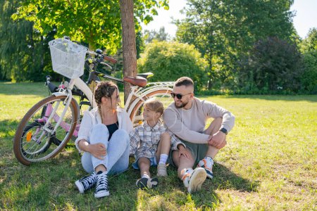 Téléchargez les photos : Famille avec enfant après le voyage à vélo, assis sur l'herbe et au repos, amusez-vous et passer du temps ensemble - en image libre de droit