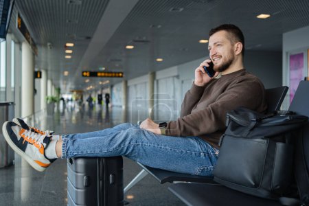 Foto de Retrato de hombre alegre relajándose en el aeropuerto con bolsa y teléfono móvil, chico esperando el vuelo utilizando el teléfono móvil que se comunica en las redes sociales, buscando información en línea. - Imagen libre de derechos