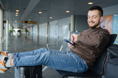 Foto de Retrato de hombre alegre relajándose en el aeropuerto con bolsa y teléfono móvil, chico esperando el vuelo utilizando el teléfono móvil que se comunica en las redes sociales, buscando información en línea. - Imagen libre de derechos