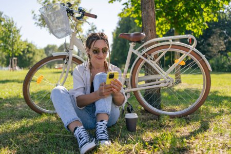 Photo for Smiling young hipster woman with bicycle using mobile phone in city park - Royalty Free Image