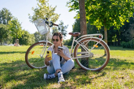 Téléchargez les photos : Smiling young hipster woman with bicycle using mobile phone in city park - en image libre de droit