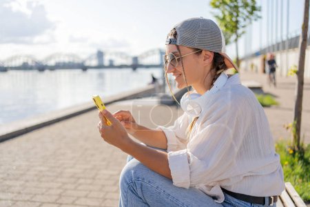 Photo for Young Hispanic hipster girl browsing mobile phone while sitting in street with takeaway beverage. - Royalty Free Image