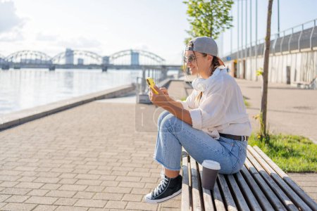 Photo for Young Hispanic hipster girl browsing mobile phone while sitting in street with takeaway beverage. - Royalty Free Image