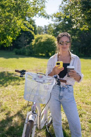 Téléchargez les photos : Smiling young hipster woman with bicycle using mobile phone in city park - en image libre de droit