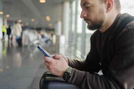 Foto de Un turista ocupado localizando en la sala de espera. Joven con celular en el aeropuerto mientras espera el avión - Imagen libre de derechos