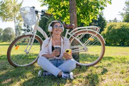 Photo for Close up of happy young girl listening to music with mobile phone and earphones while sitting at the park with a bicycle. Lifestyle. Relax, nature concept - Royalty Free Image