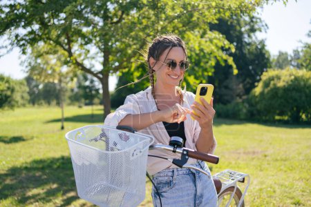 Photo for Smiling young hipster woman with bicycle using mobile phone in city park. Talking by video call, using internet - Royalty Free Image
