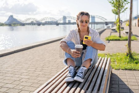 Téléchargez les photos : Femme hipster avec tresses et café à emporter assis à l'aide d'un smartphone dans la rue de la ville. Heureuse étudiante assise à l'extérieur avec smartphone dans les mains pendant la pause café avoir un appel vidéo avec des amis. - en image libre de droit