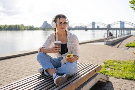 Photo for Hipster woman in bluetooth headphones enjoying positive music playlist from mobile radio using 4g wireless in city, have video call, girl holding coffee to go and smartphone. - Royalty Free Image