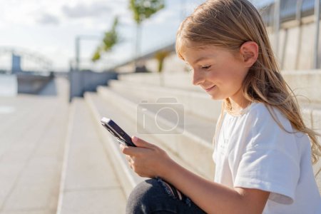 Téléchargez les photos : Enfant fille assis à la rue en ville utiliser un téléphone portable. - en image libre de droit