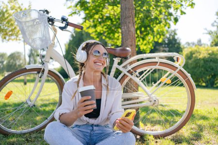 Foto de Primer plano de la joven feliz escuchando música con teléfono móvil y auriculares mientras se sienta en el parque con una bicicleta. Estilo de vida. Relájate, concepto de naturaleza - Imagen libre de derechos