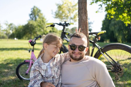Téléchargez les photos : Père riant et sa fille s'amusent ensemble au parc vert, se détendre et s'amuser ensemble après avoir fait du vélo . - en image libre de droit
