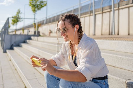 Photo for Smiling beautiful woman holding mobile phone listening music, watching video on the street. Happy hipster female using mobile app shopping online outdoors. - Royalty Free Image