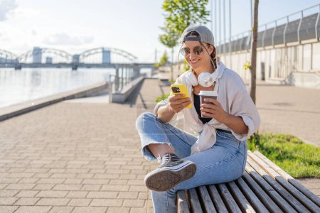 Téléchargez les photos : Jeune hispanique hipster fille navigation téléphone mobile tout en étant assis dans la rue avec boisson à emporter. - en image libre de droit