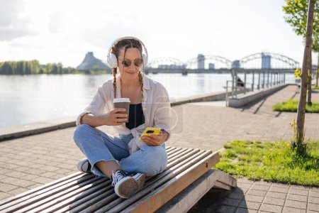 Photo for Hipster woman in bluetooth headphones enjoying positive music playlist from mobile radio using 4g wireless in city, have video call, girl holding coffee to go and smartphone. - Royalty Free Image