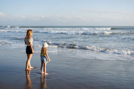 Téléchargez les photos : Mère avec sa fille s'amuser à la plage avec du sable noir à Bali, courir sur les vagues, profiter de la vie et voyager - en image libre de droit