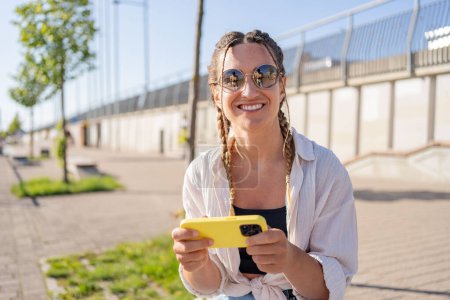 Photo for Positive hipster girl holds mobile phone, looks at cellphone screen, uses smartphone apps. girl with mobile phone on the street. - Royalty Free Image