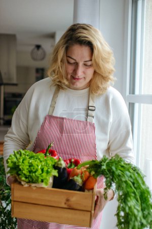 Foto de Una mujer sosteniendo una caja de madera llena de verduras frescas crudas. Cesta con verduras zanahorias, pimientos, berenjenas, ensalada en sus manos. - Imagen libre de derechos