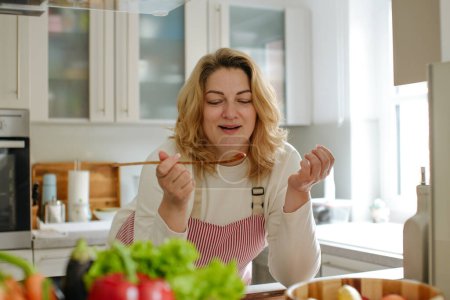 Photo for Young woman eating salad at table with organic vegetables, enjoying healthy diet, standing in light kitchen interior. Lady cooked veggie meal at home. Weight loss concept - Royalty Free Image