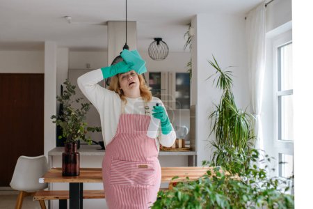 Photo for A woman with blonde hair wearing gloves and carrying a rag is cleaning a bright flat with flowers - Royalty Free Image