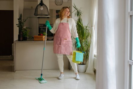 Foto de Mujer sonriente mirando por la ventana y limpiando la cocina - Imagen libre de derechos