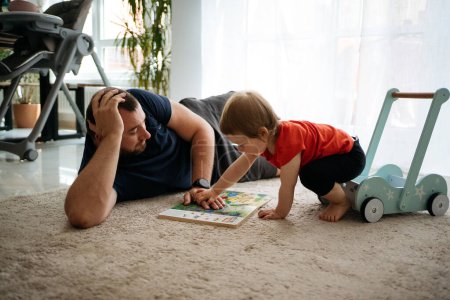 Téléchargez les photos : Un père et sa jeune fille, portant un t-shirt rouge, s'assoient sur le sol d'un appartement spacieux et regardent un livre pour enfants - en image libre de droit