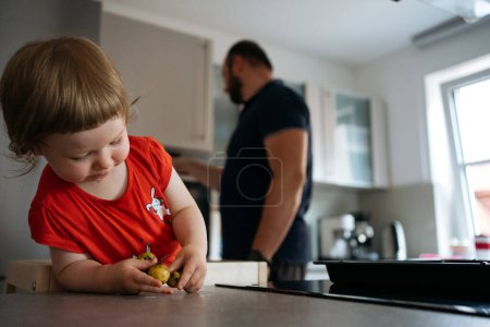 Téléchargez les photos : Une petite fille est dans la cuisine à jouer avec les fruits pendant que son père prépare la nourriture - en image libre de droit