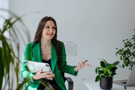 Téléchargez les photos : Portrait de ceinture de femme d'affaires moderne au bureau avec tablette graphique. Femme en vêtements de travail veste verte assis avec bureau léger avec des fleurs - en image libre de droit