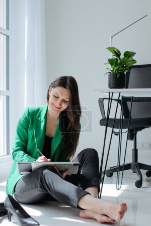 Foto de Una joven está sentada en el suelo en una oficina, con una tableta gráfica en las manos. Ella se ha quitado los zapatos y sonríe - Imagen libre de derechos
