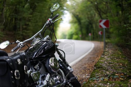 Foto de Una motocicleta negra de estilo clásico se encuentra al lado de una carretera sinuosa rodeada de bosque - Imagen libre de derechos