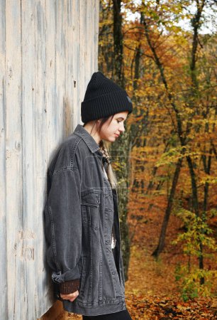 Foto de Una chica morena con un sombrero y una chaqueta de mezclilla gris está de costado cerca de una casa de madera en el contexto de un bosque de otoño. - Imagen libre de derechos