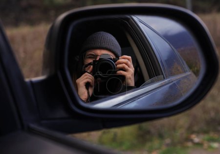 Foto de Una chica morena con gafas mira al visor de la cámara y se refleja en el espejo del coche en un sombrío día de otoño. - Imagen libre de derechos