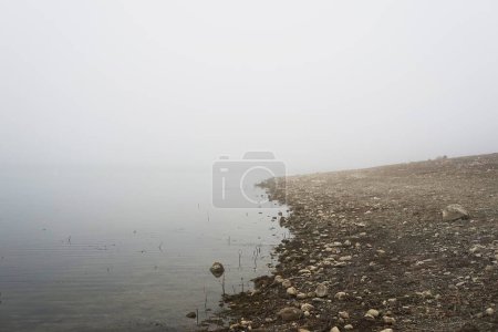 Foto de Orilla rocosa cerca de la superficie tranquila del lago en tiempo de niebla - Imagen libre de derechos