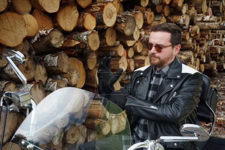 Foto de Un hombre con gafas y una chaqueta de cuero se sienta en una motocicleta y se pone guantes contra el fondo de los cortes de árboles en un día sombrío - Imagen libre de derechos