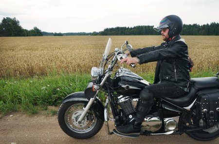 Foto de Un hombre en un casco en una motocicleta negra clásica en el fondo de un campo en un día nublado de verano - Imagen libre de derechos