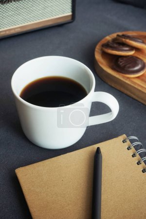 Foto de Taza blanca con café junto a galletas en una bandeja de madera, un bloc de notas y una columna de música en una mesa gris - Imagen libre de derechos