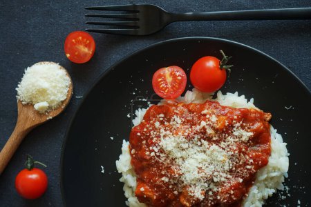 Foto de Arroz con salsa de tomate, albóndigas y parmesano en un plato negro junto a un tenedor y tomates cherry sobre un mantel gris - Imagen libre de derechos